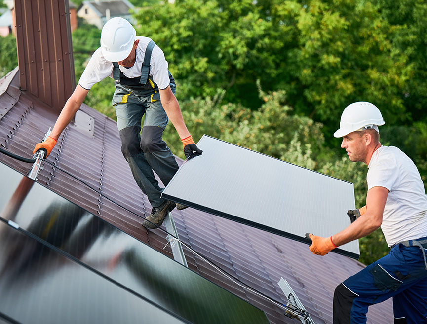 Two people wearing hard hats and work clothes are installing a solar panel on a metal roof. One person is on the lower part of the roof holding the panel, while the other is on the upper part, securing it.